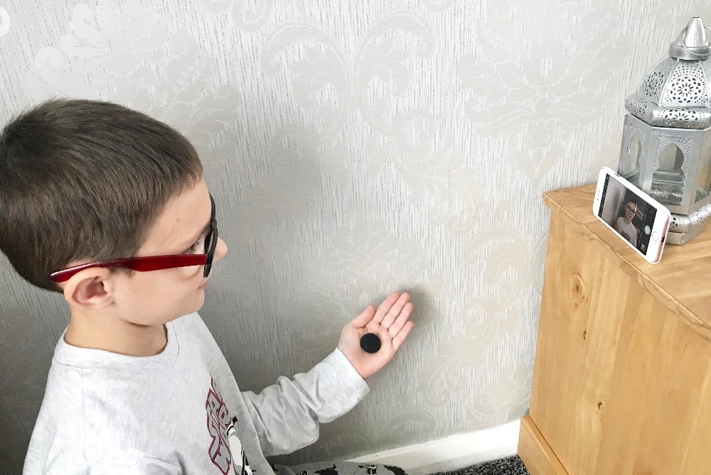 A boy wearing glasses holds a small round Flic button in his hand and looks at a mobile propped up on a wooden surface, with a decorative lantern beside it, hinting at smart home automation.