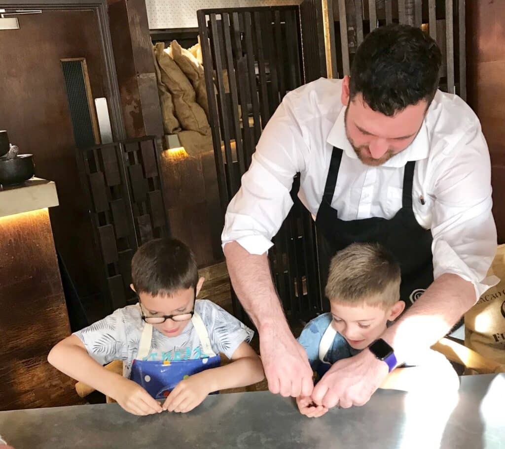 A man wearing a black apron helps two young boys with a cooking activity at a table in the cosy School Of Chocolate. The boys, also in aprons, focus on their hands as they work with small items, sunlight streaming into the room.