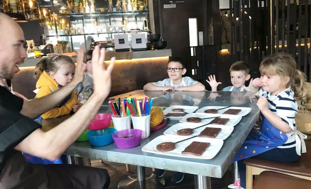 A chef from the School of Chocolate leads a chocolate-making activity for five young children seated at a table with moulds, bowls of toppings, and colourful markers in a bright Leeds kitchen. The children are raising their hands and smiling.