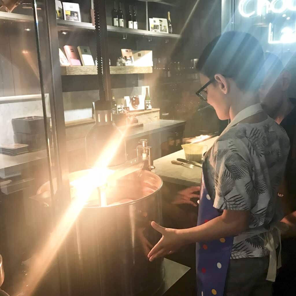 A young boy wearing glasses and a colourful apron stands by a large metal vat at the Hotel Chocolat School of Chocolate in Leeds, watching chocolate being prepared in a well-lit kitchen with shelves of bottles and boxes in the background.