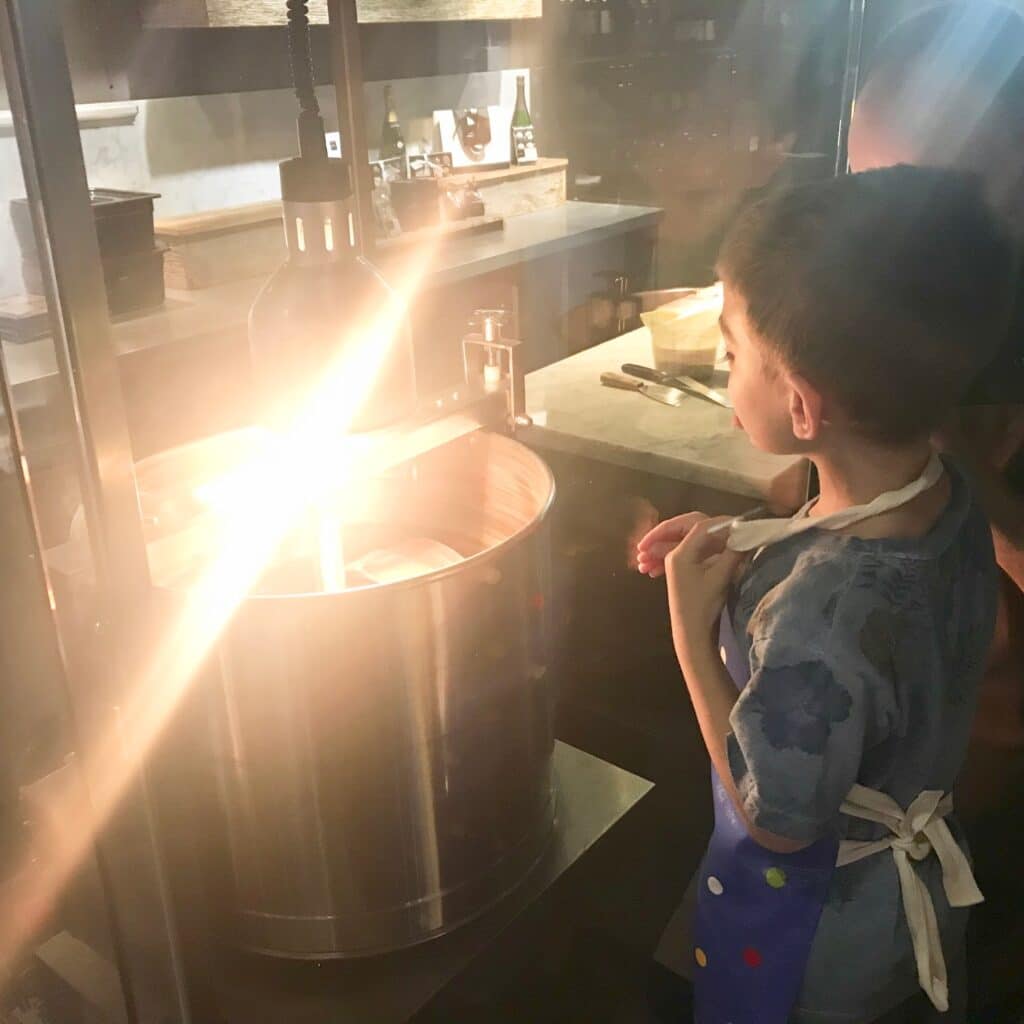 A young boy wearing an apron stands closely watching a candyfloss machine spin under a bright light in a modern Leeds kitchen, reminiscent of a School Of Chocolate workshop.