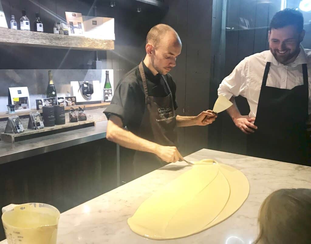 A chef spreads a large sheet of yellow batter or chocolate on a marble worktop with a spatula, while another person in an apron looks on and smiles in a modern kitchen—perfect for aspiring chocolatiers at School Of Chocolate.