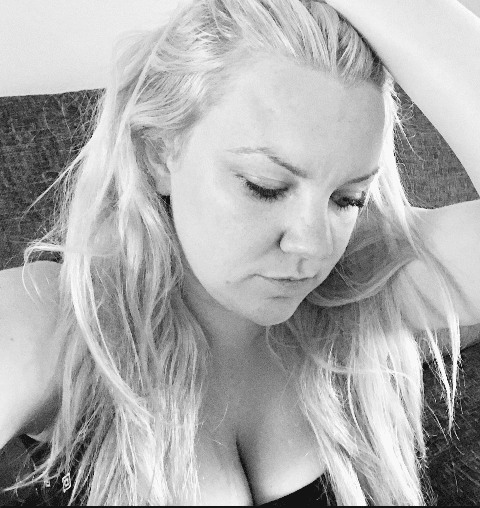 A woman with long, fair hair sits on a sofa, looking downward with a thoughtful expression. Her hand rests on her head as she reflects on motherhood and body image. The image is captured in striking black and white.