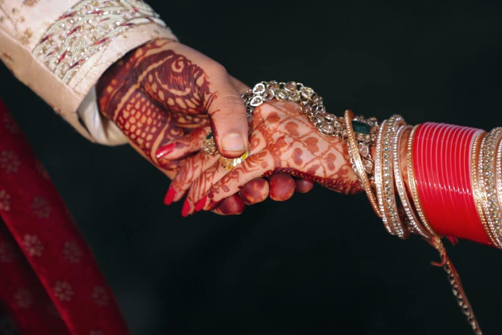 A close-up of two hands clasped together, adorned with intricate henna designs, bangles, and jewellery, symbolising an Indian wedding or engagement ceremony and showcasing a beautiful outfit for a wedding of a different culture.