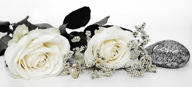 Two white roses with small white flowers, two gold rings, and a grey stone with "Love" written on it are arranged on a white surface—perfect inspiration for an outfit for a wedding of a different culture.