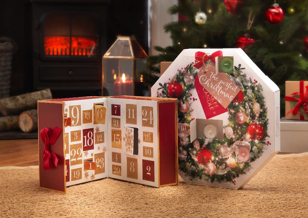 A festive room with a lit fireplace, stacked logs, and a decorated Christmas tree. Two family-friendly Christmas Advent Calendars—one open showing numbered doors—sit on the floor beside a wreath-themed round calendar.