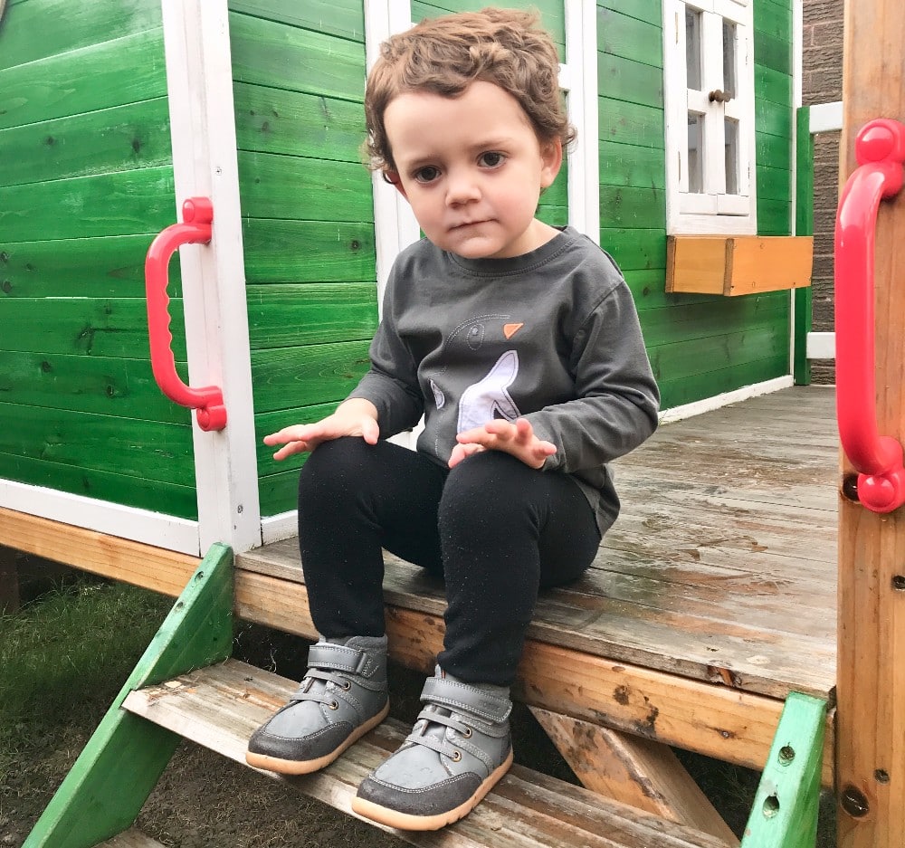 A young child with curly hair sits on wooden steps outside a green playhouse, wearing a grey long-sleeve shirt, black trousers, and cosy Bobux footwear—perfect for autumn—looking slightly at the camera with hands resting on their knees.