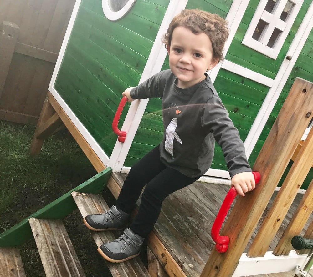 A young child with curly hair smiles whilst standing on the steps of a green wooden playhouse, wearing Bobux footwear and holding onto red handles on each side of the stairs.