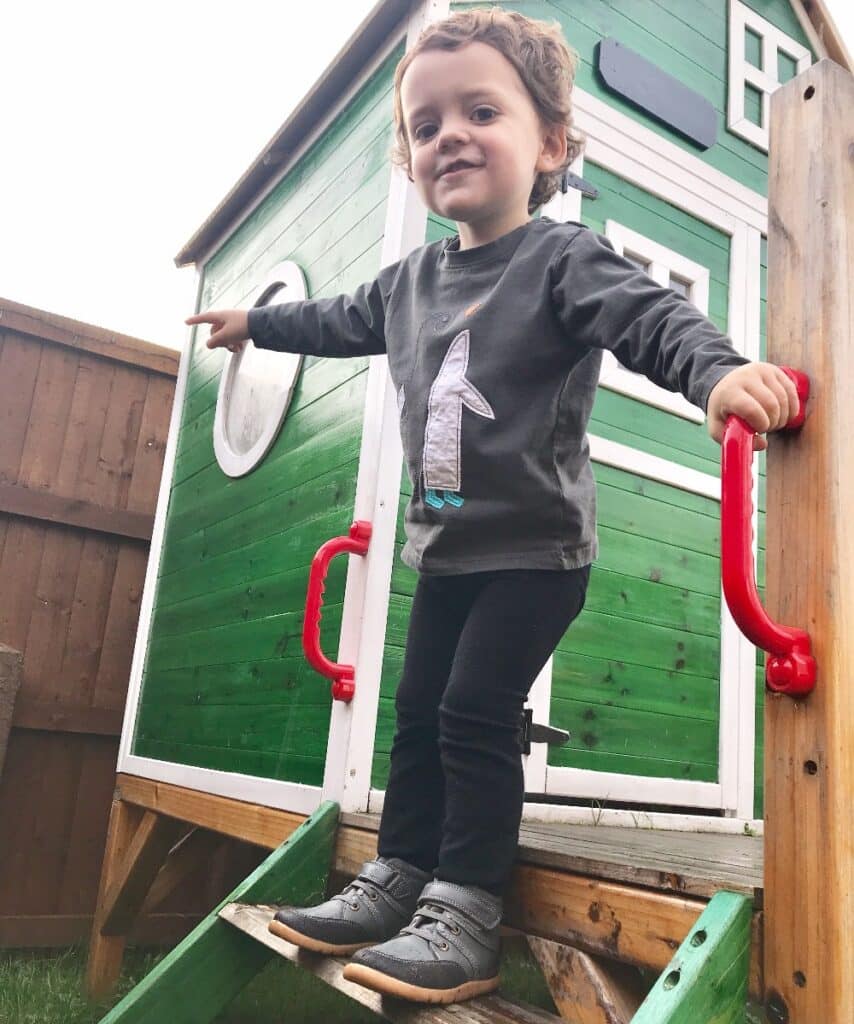 A young child in Bobux Footwear stands on steps leading to a small, green wooden playhouse with a circular window, holding onto red handrails and smiling whilst pointing outward.