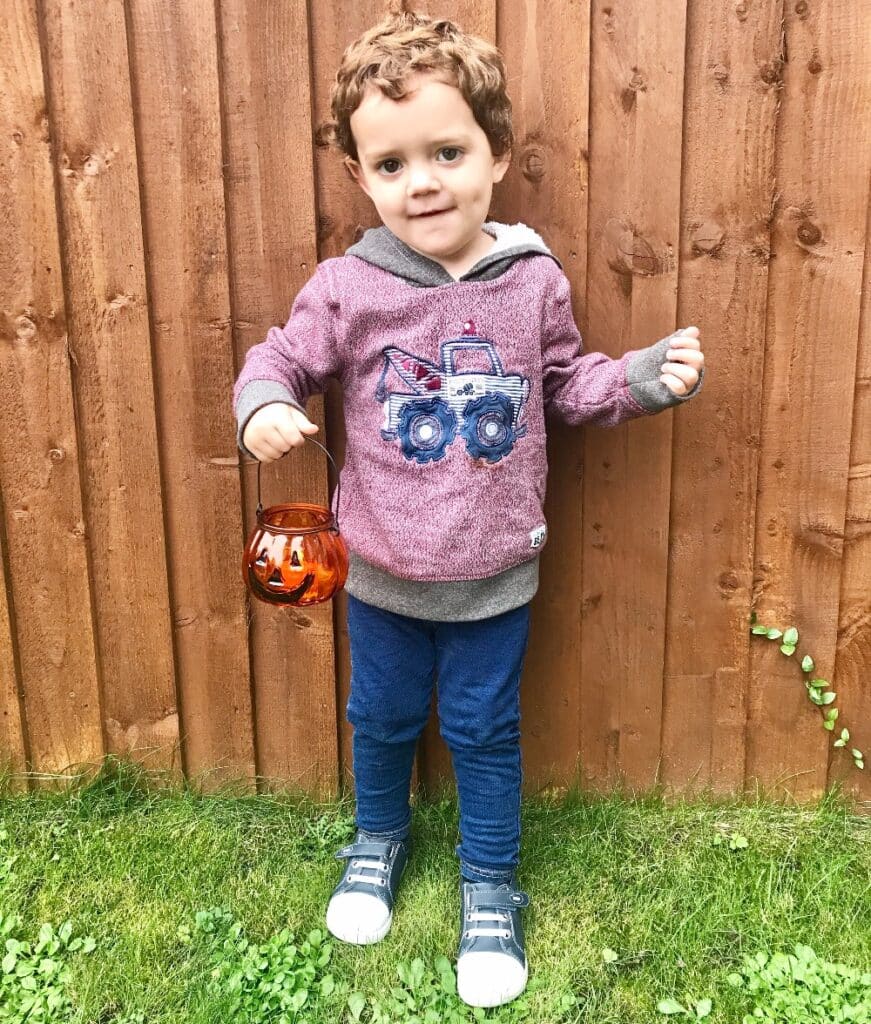 A young boy with curly hair stands on grass in front of a wooden fence, wearing a hoodie with a tractor design, blue trousers, and grey Bobux footwear. He holds an orange pumpkin-shaped lantern, ready for a crisp autumn evening.