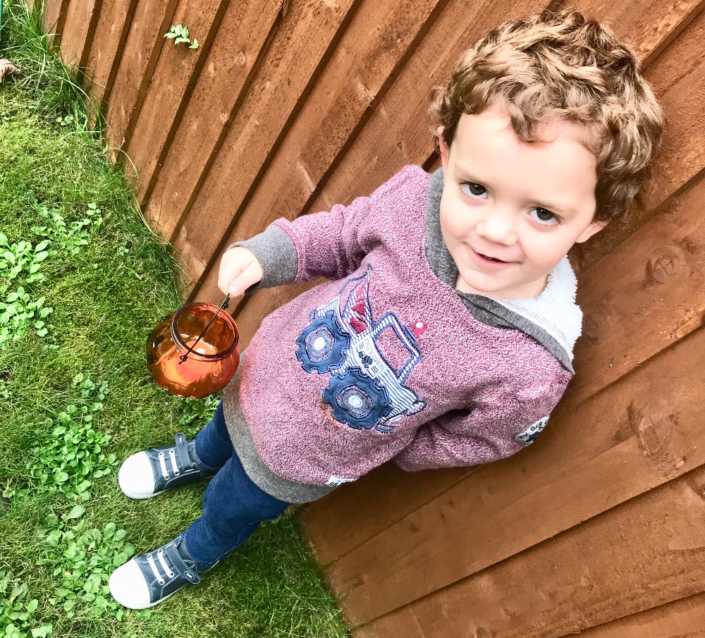 A young child with curly hair, wearing a maroon jumper with a monster truck design, blue jeans, and blue Bobux footwear, stands on autumn grass next to a wooden fence, holding a small brown glass jar.