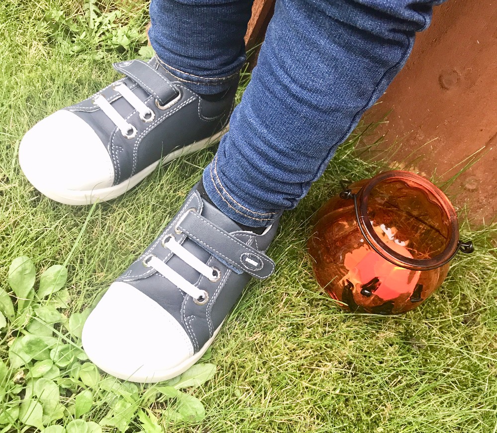 A child wearing blue jeans and navy Bobux footwear stands on grass next to an amber-coloured glass jar, capturing a cosy autumn vibe.