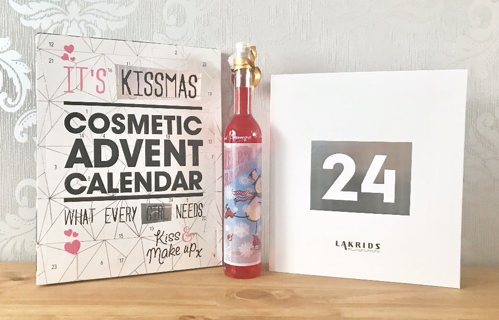 Three holiday-themed items on a table: a “Cosmetic Advent Calendar,” a tall bottle with a gold bow, and a white box labelled “24 LAKRIDS”—perfect Advent Calendars to share with family, set against a light patterned background.