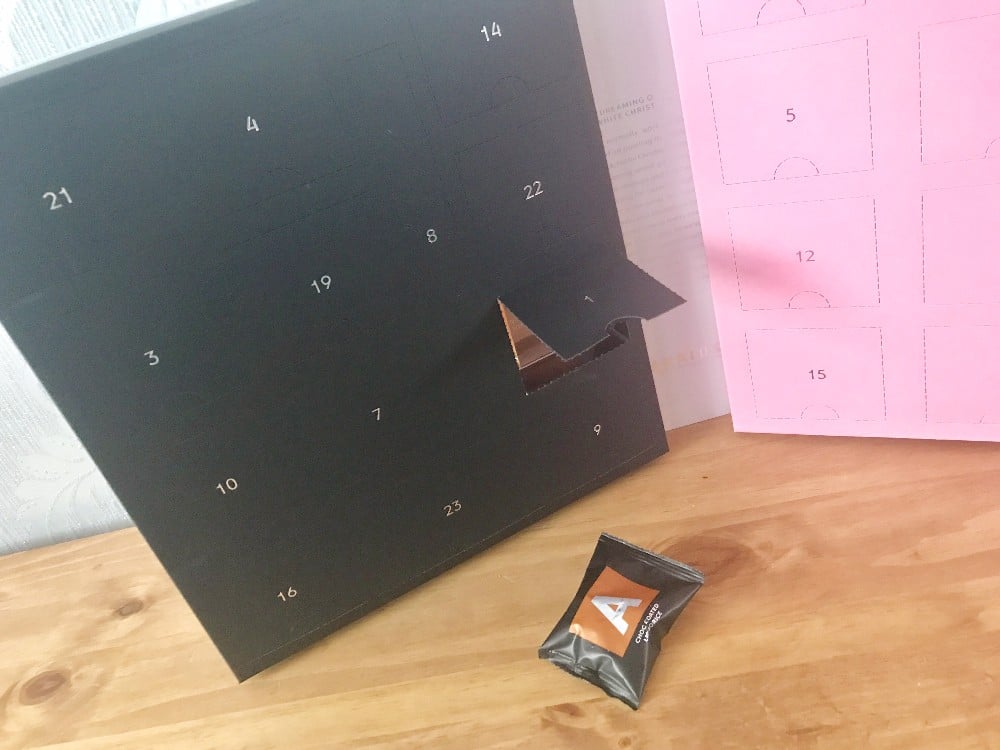 A black Advent Calendar with one door open sits on a wooden surface. In front is a small, black-wrapped item labelled with a white "A". A pink sheet with numbered boxes hints at the fun of Christmas Advent Calendars for the whole family.