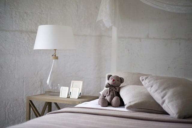 A cosy bedroom makeover featuring a bed with beige bedding, two pillows, and a brown chequered teddy bear. A bedside table holds a white lamp and two small picture frames against a white textured wall, creating stylish bedroom decor.