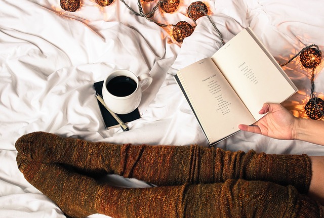 Person wearing snug brown socks sits on a bed with white sheets, holding an open book. A cup of coffee and a notebook with a pen are beside them, along with decorative fairy lights, creating a cosy space perfect for relaxing.