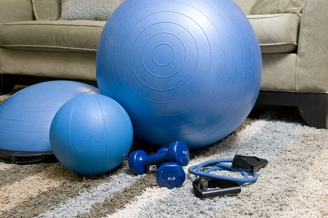 A set of blue exercise equipment, ideal to create a home gym, including a stability ball, medicine ball, BOSU ball, dumbbells, and resistance band, rests on a carpeted floor in front of a beige sofa.