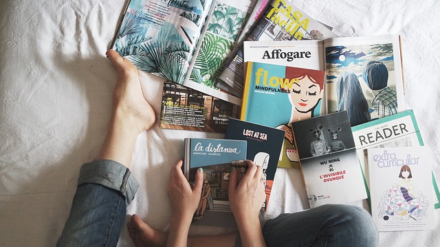 Person sitting on a bed with bare feet, surrounded by various open books and magazines, holding a book titled "La Distanza." The scene suggests relaxation in a cosy space perfect for unwinding and enjoying some quiet reading time.