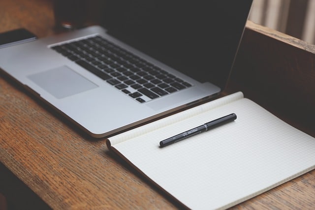 A silver laptop sits on a wooden desk next to an open, blank squared notebook with a black pen resting on it, perfect for jotting down family extension planning ideas or making space for new possibilities.