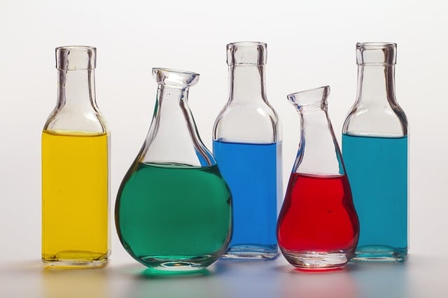 Five glass bottles filled with colourful liquids—yellow, green, blue, red, and teal—are arranged in a row against a light background. The varied bottles highlight how buying in bulk can help save money and reduce household waste.
