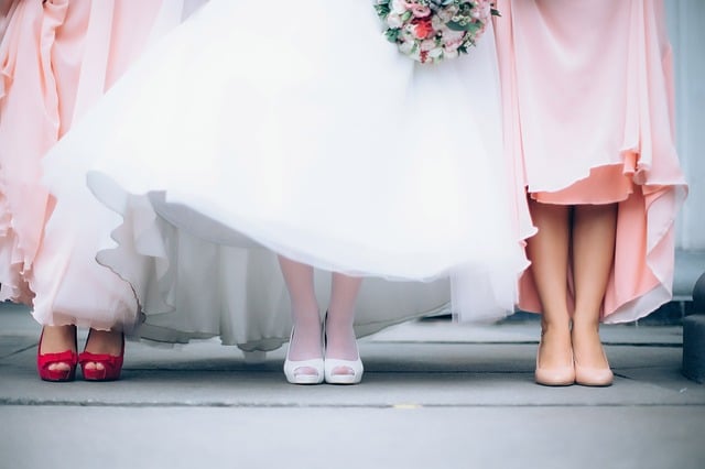 Three women stand side by side in formal dresses; the centre wears a white gown and holds a pink bouquet, while her budget bridesmaids wear blush dresses with pink and red heels. Faces are not visible.