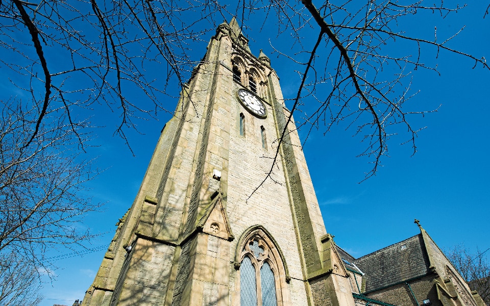 A tall stone church tower with a large clock, viewed from below against a clear blue sky. Bare tree branches frame the foreground, adding striking definition to the scene.