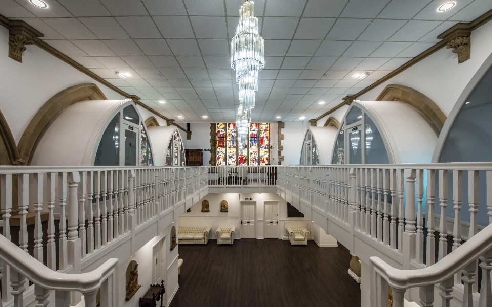 Spacious, elegant interior with white railings, arched doorways, a grand chandelier, stained glass windows at the far end for stunning effect, and beige sofas along the walls on a dark wooden floor.