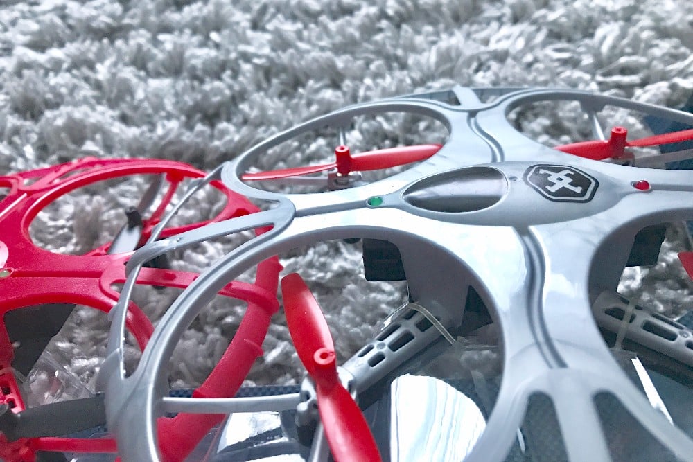 Close-up of two Air Wars Battle Drones with plastic propeller guards, one grey and one red, resting on a textured white carpet. The grey drone is in the foreground, showing its central logo and propellers in detail.