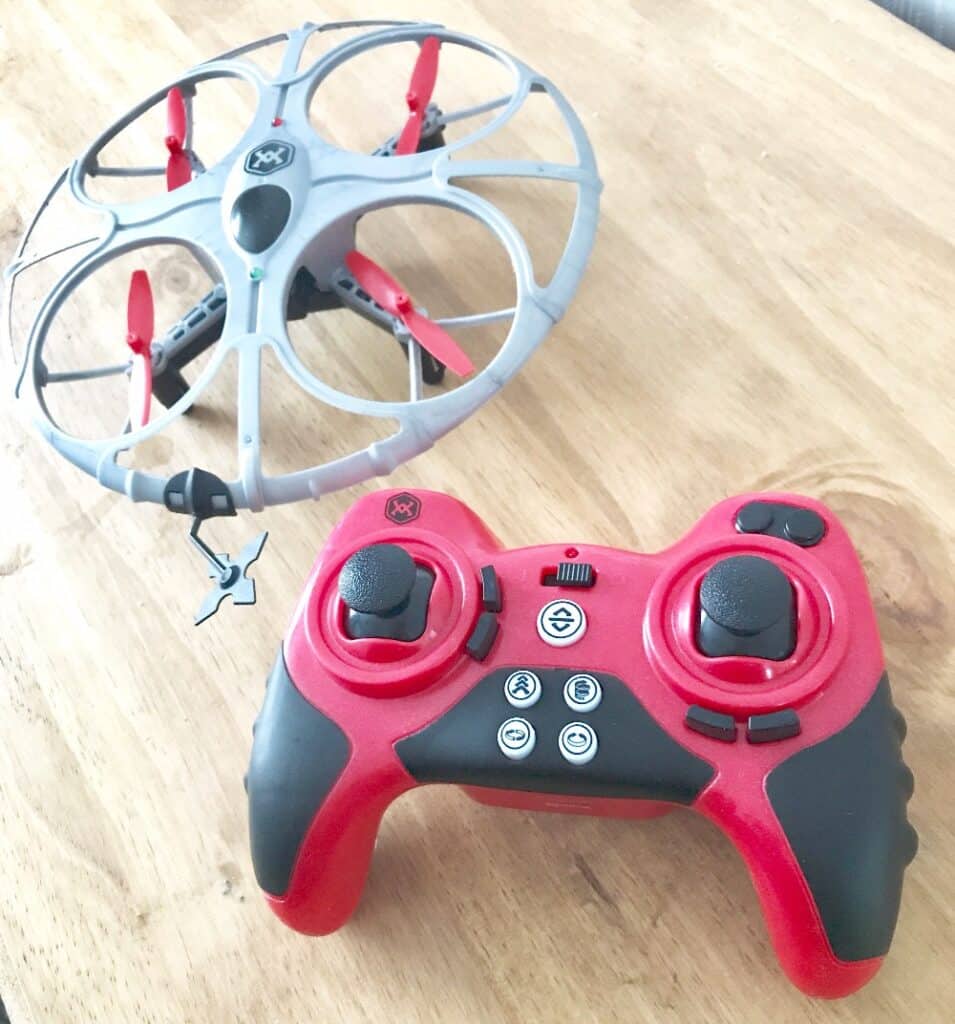 A small red and grey Battle Drone sits on a wooden surface next to a matching red and black remote control with two joysticks and several buttons, ready for thrilling Air Wars action.