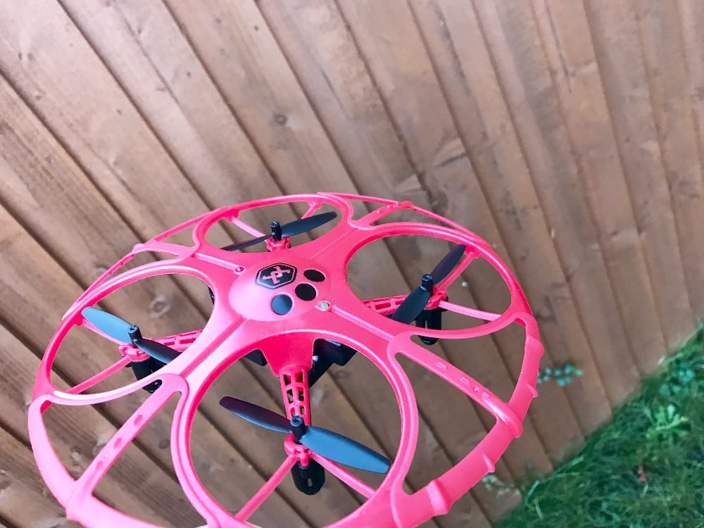 A red Air Wars quadcopter drone with propeller guards is flying close to a wooden fence, with a patch of green grass visible in the lower right corner—perfect for your next Battle Drones review.