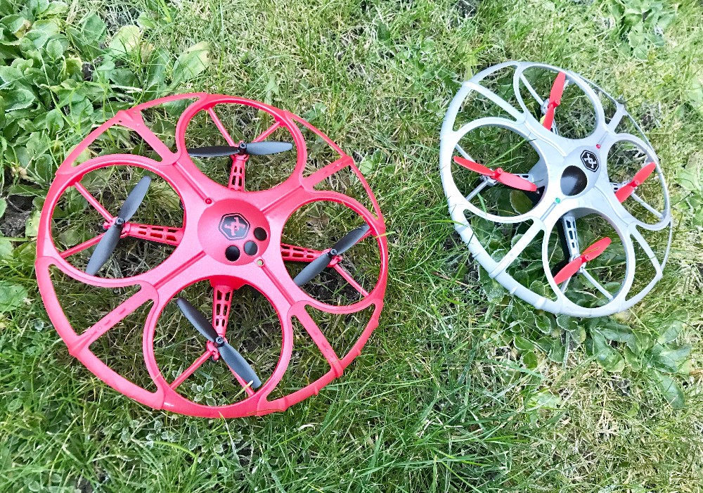 Two round Battle Drones with protective outer rings—one red, one grey—rest on green grass. Both feature four propellers and a central camera or sensor area, ready for an exciting Air Wars showdown.