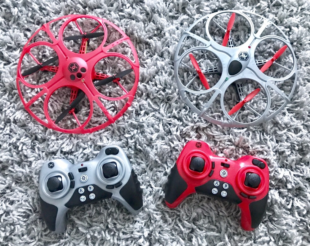Two small Air Wars Battle Drones, one red and one silver, with matching controllers rest on a textured grey carpet. The circular drones feature protective frames around the blades, perfect for thrilling Air Wars Drones matches at home.