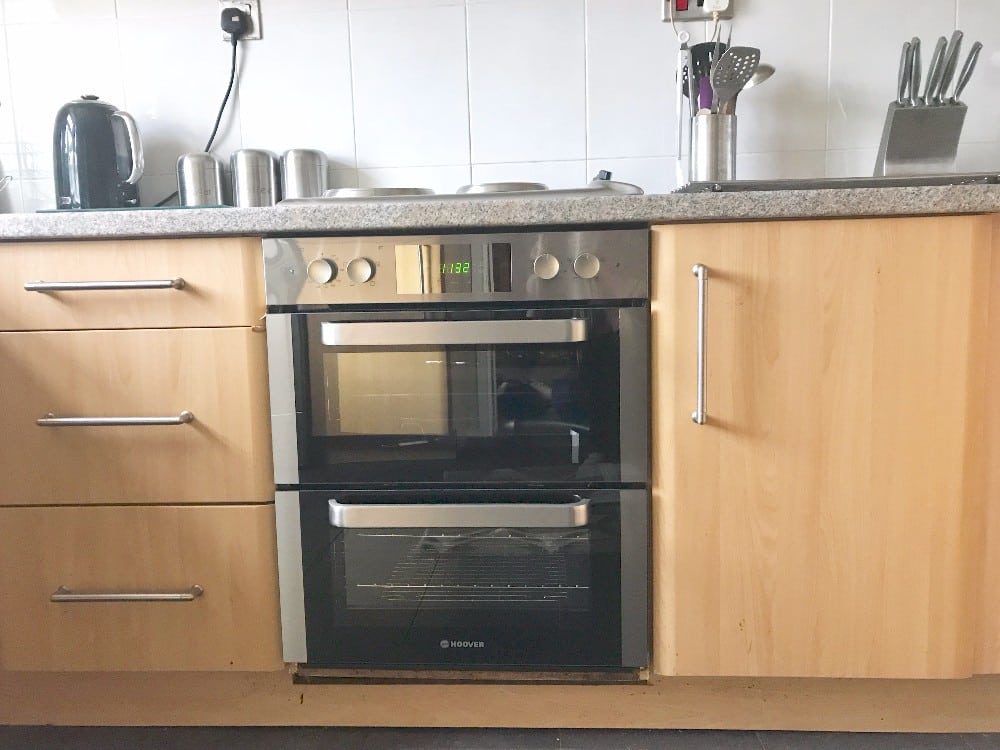 A modern Hoover HO7D3120IN double oven is built into a wooden kitchen unit. The worktop holds a kettle, metal canisters, and a utensil holder with kitchen tools. A knife block sits on the right. Available at AO.com.