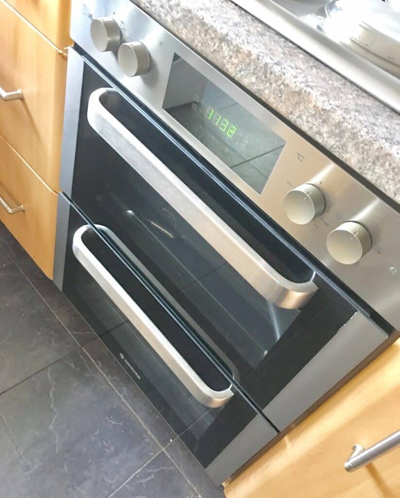 A modern Hoover double oven, model HO7D3120IN, with stainless steel and black finish, digital clock at 11:32, and silver control knobs, set in a kitchen with wooden cupboards and a dark tiled floor. Available at AO.com.