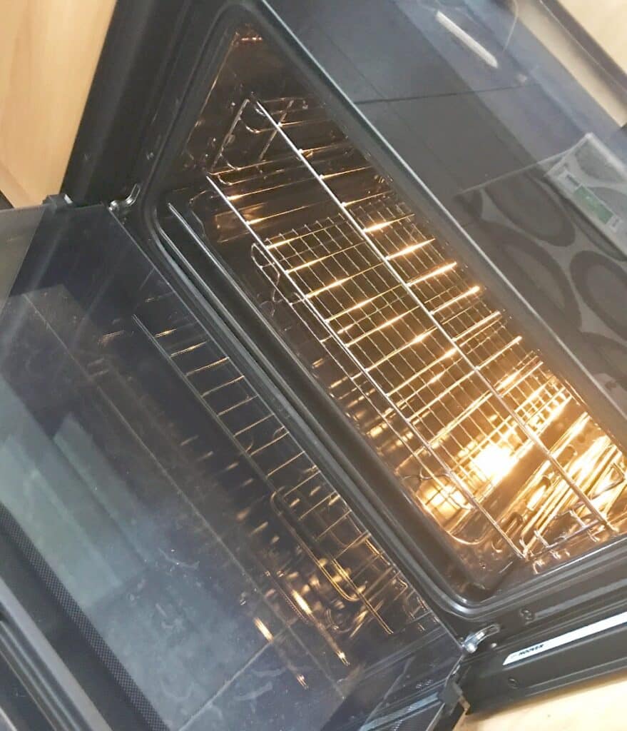 An open Hoover HO7D3120IN double oven with the door pulled down and the interior light on, revealing empty metal shelves inside. The clean oven appears to be built into a kitchen cupboard, as featured on AO.com.