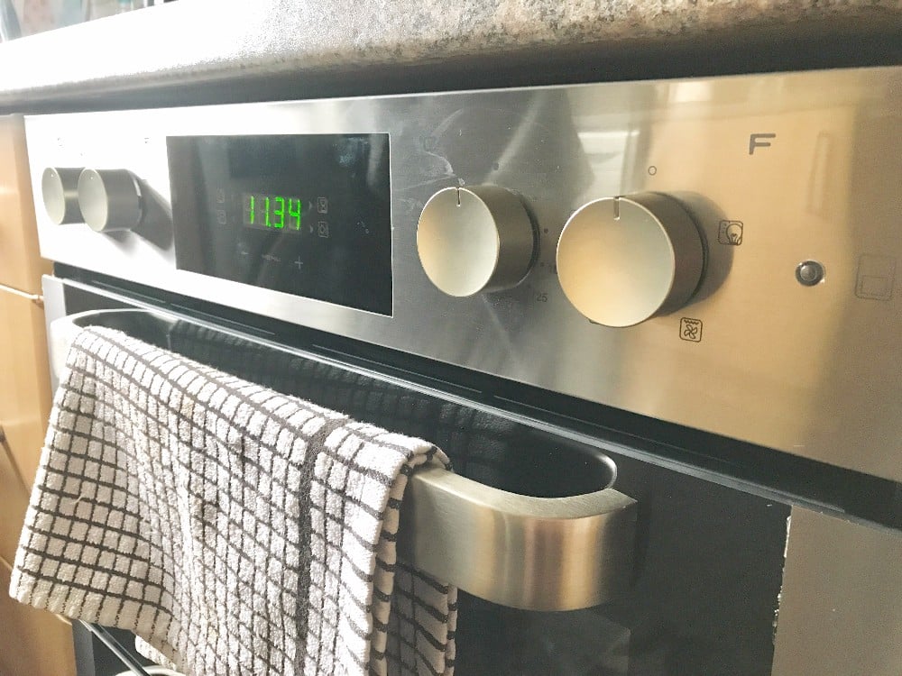 A Hoover HO7D3120IN stainless steel double oven with a digital clock displaying 11:34, three control knobs, and a chequered tea towel hanging on the oven door handle—available now at AO.com.