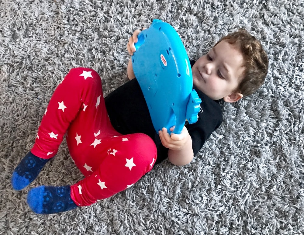 A young child wearing red star-patterned trousers and blue socks lies on a grey carpet, holding and looking at a large Thomas & Friends tablet toy.