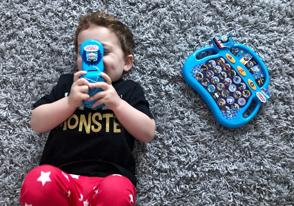 A young child in red star-patterned trousers and a black "monster" shirt lies on a grey carpet, smiling and holding a Thomas & Friends Flip & Learn Phone. A blue Alpha-Phonics educational toy tablet with buttons lies nearby.