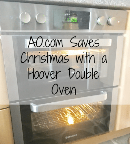 A stainless steel Hoover HO7D3120IN double oven with both doors closed and interior lights on. Overlaid text reads: “AO.com Saves Christmas with a Hoover Double Oven.” The oven is built into a kitchen unit.