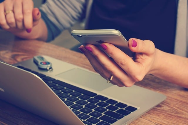 A new graduate with pink nail varnish holds a mobile phone in one hand while using a laptop on a wooden desk—classic multitasking for an entrepreneur starting up. A USB memory stick and another hand resting on the laptop are also visible.