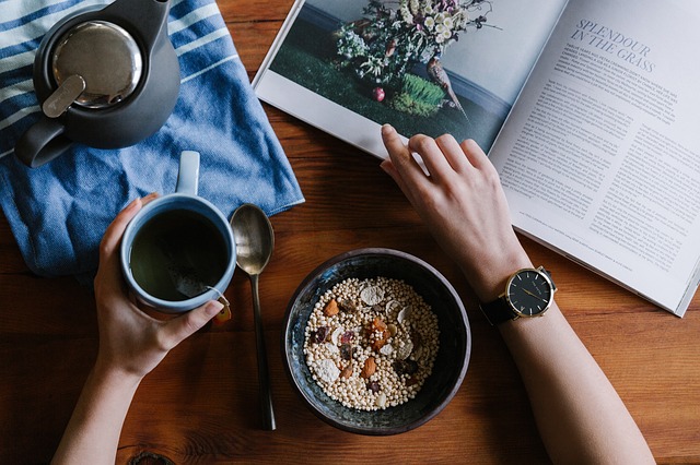 A person sits at a wooden table with a bowl of cereal and nuts, a cup of tea, a teapot, and an open magazine—finding quiet moments to cope with depression as one hand holds the mug and the other rests on the magazine.