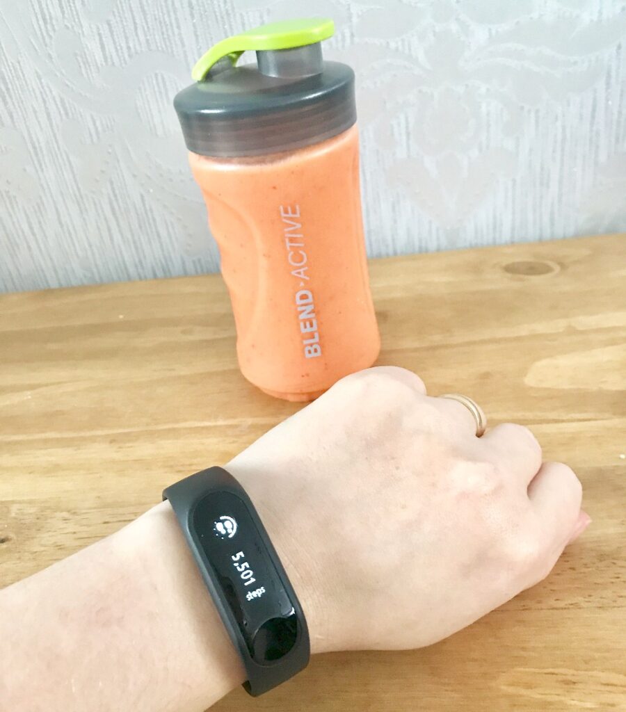 A hand wearing a black fitness tracker displaying 5,507 steps rests on a wooden surface. In the background, a Blend Active bottle filled with an orange smoothie hints at motivation towards reaching 10,000 steps for Teenage Cancer Trust.