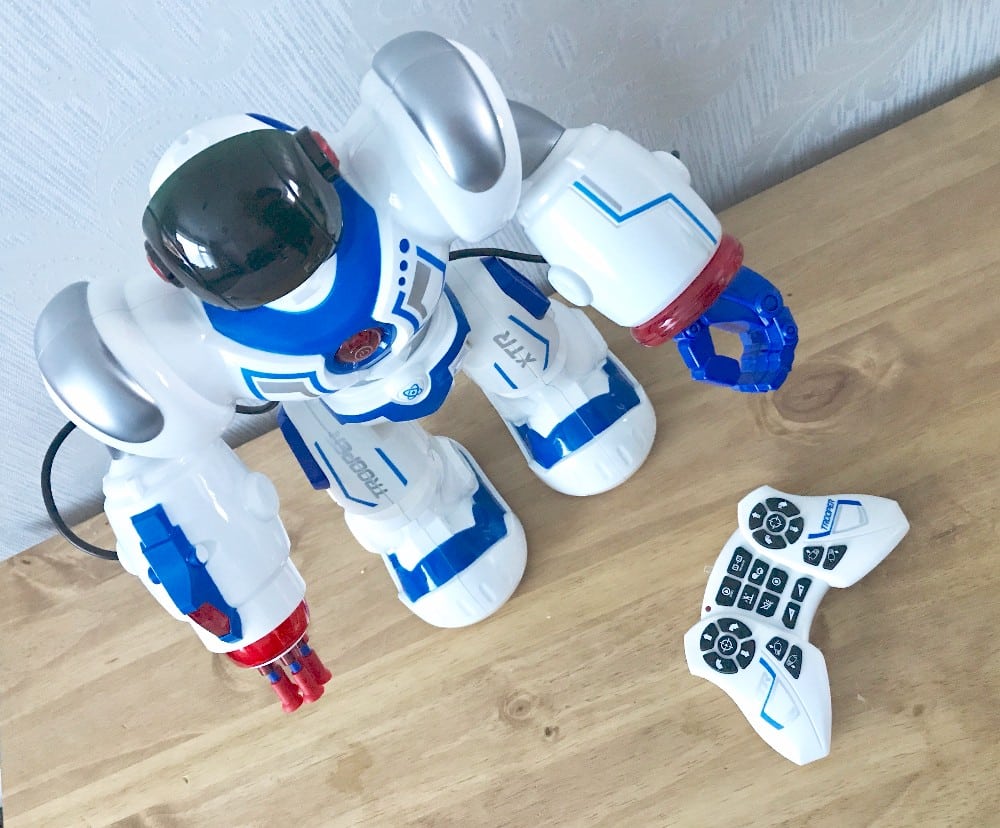 A white and blue XT30039 Trooper Bot toy robot with a black visor stands on a wooden surface next to a matching Xtrem Bots remote control. The robot’s arm is raised, set against a light-coloured wall background.