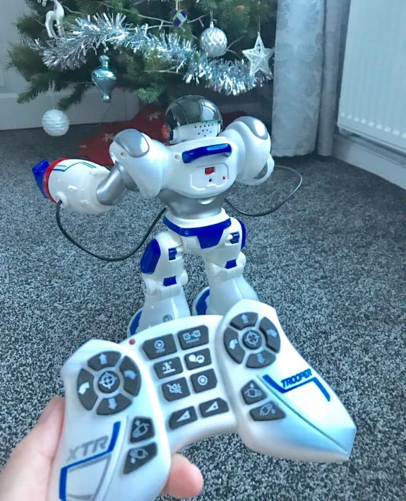 A person holds a white and blue Xtrem Bots remote control aimed at the Trooper Bot robot on the floor in front of a decorated Christmas tree. The robot features a helmet and blue accents, model XT30039.