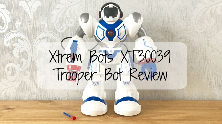 A white and blue Xtrem Bots Trooper Bot (XT30039) stands on a wooden surface before a decorative wall. Text over the image reads: "Xtrem Bots XT30039 Trooper Bot Review." A small red and blue accessory lies nearby.
