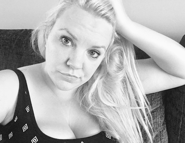 Black and white photo of a woman with long blonde hair sitting on a sofa, looking at the camera with a neutral expression. She wears a patterned top with thin straps, embodying self-acceptance and quiet body positivity.