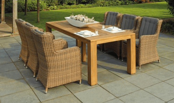 A wooden garden furniture set with six wicker chairs featuring grey cushions, set on a stone patio with grass and trees in the background. A large decorative bowl sits on this stylish piece of outdoor furniture.