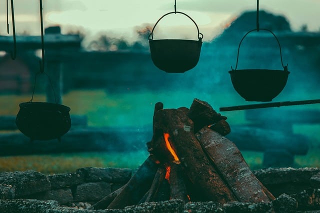 Three black pots hang over a campfire with large logs, surrounded by a stone ring. Blue smoke rises as the fire burns—a classic scene from camping with a large family, whilst blurred greenery is visible in the background.