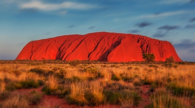 Uluru, a massive red sandstone rock formation, rises from the flat, grassy landscape under a partly cloudy sky at sunset—the perfect scene for a dream family in Australia’s outback.