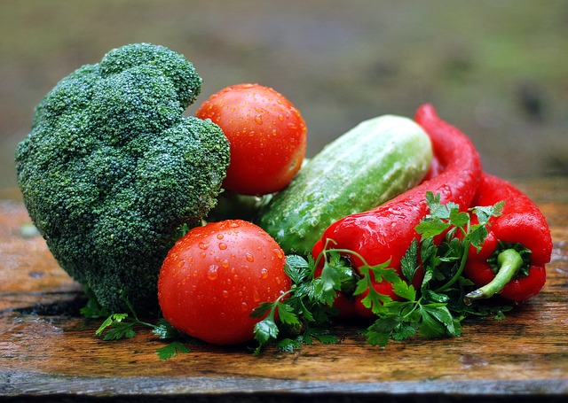A broccoli floret, two tomatoes, a cucumber, two red peppers, and parsley are arranged on a wooden surface, with water droplets visible—a fresh scene reflecting 2018 food and drink industry trends.